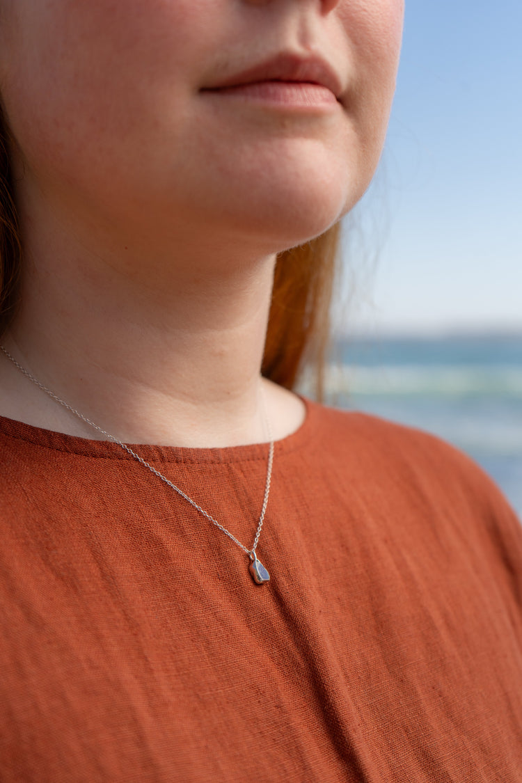 Close up of a woman in an orange linen dress wearing a blue mini sea pottery necklace with the blue sky and sea in the background