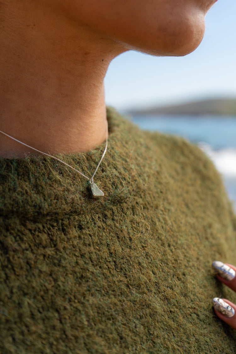 A woman in a green jumper wears a small green sea glass necklace with the sea in the background
