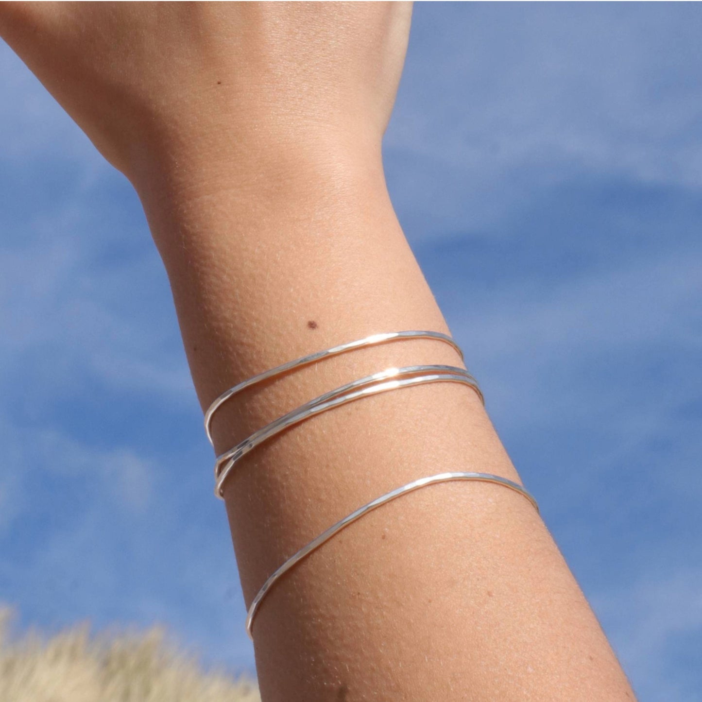 Hammered silver bangles on person's wrist with sky in background