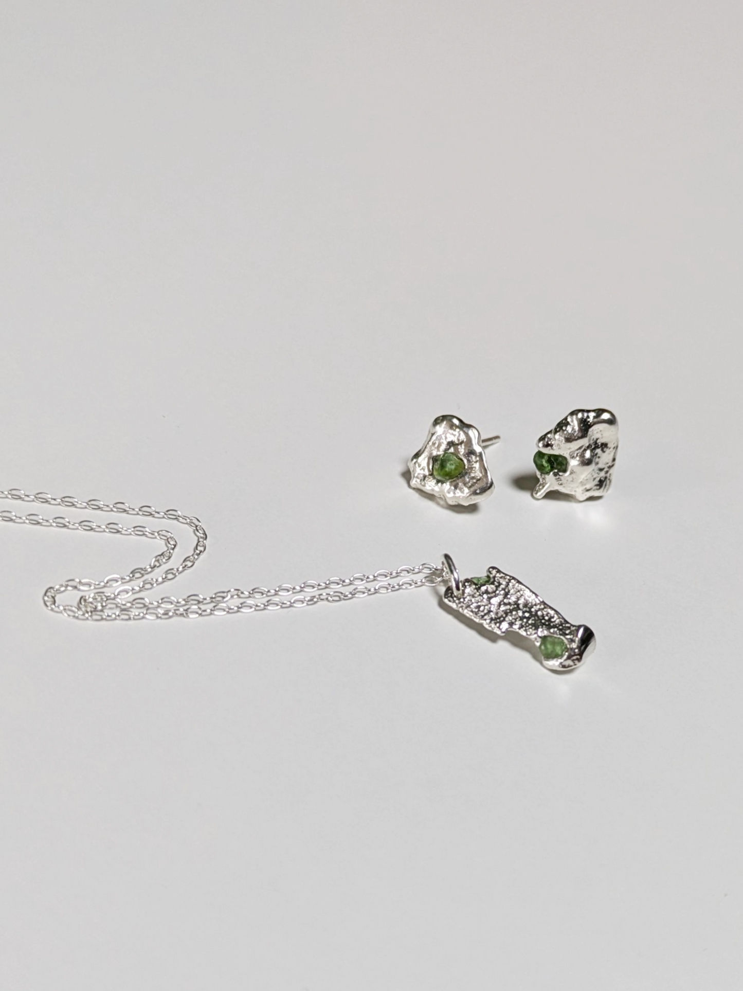 A matching pair of scrap silver earrings and necklace with green sea glass shards embedded, on a light grey background.