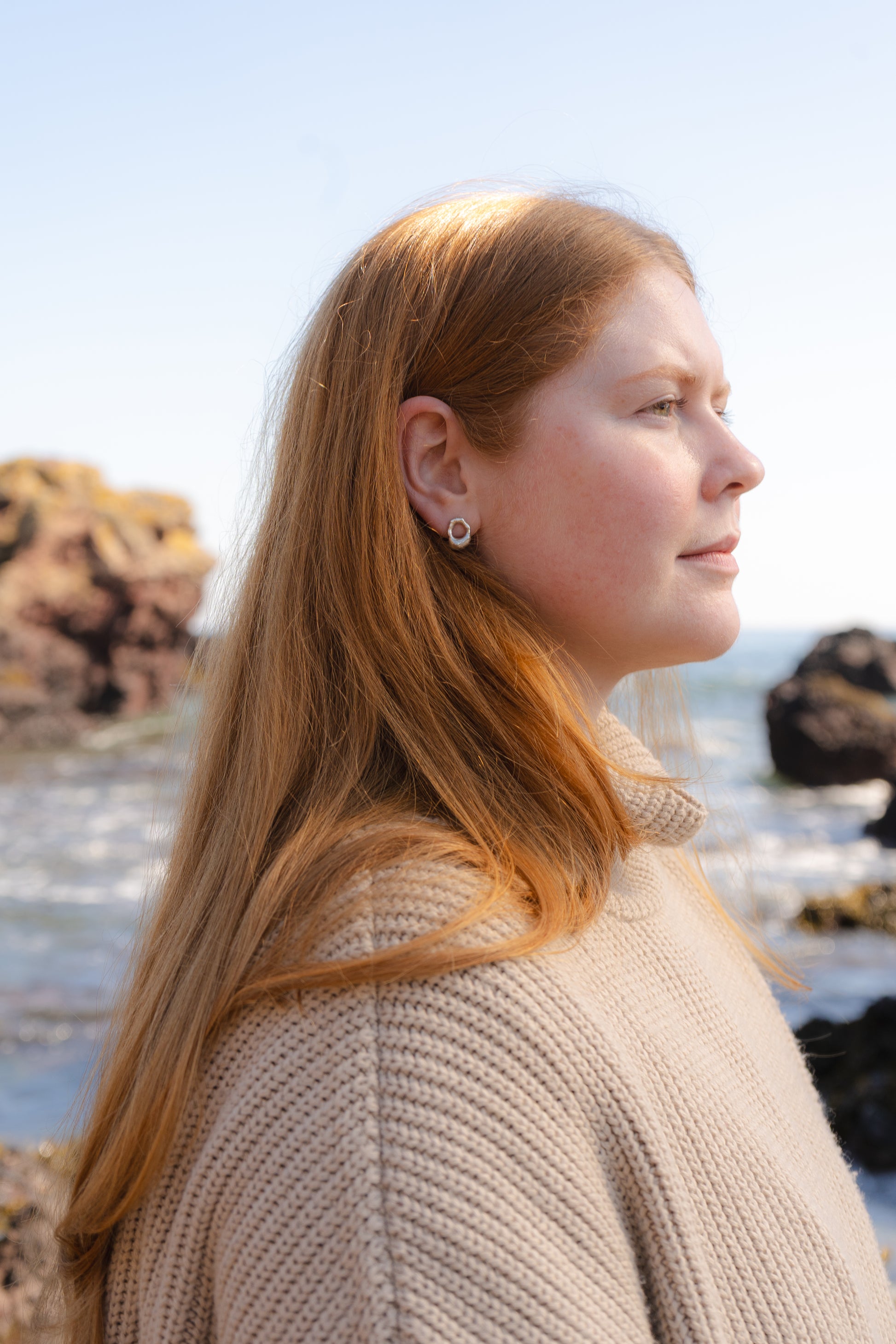 Woman with red hair standing on a beach  wearing mini molten studs, with rocks and sea in the background.