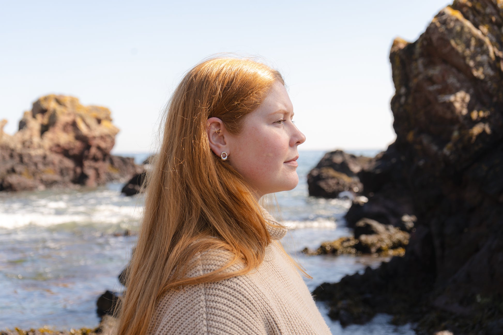 Woman with long red hair standing on a rocky beach wearing mini molten studs