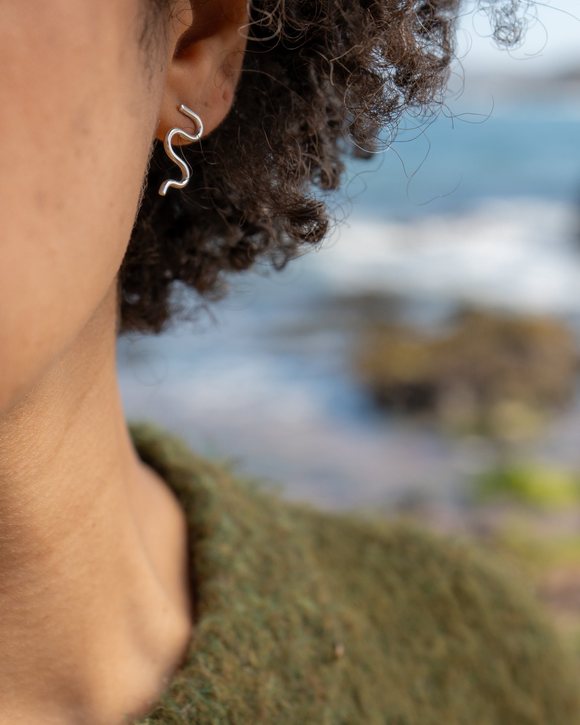Close-up of a person wearing mini wiggle earrings with a blurred natural landscape in the background