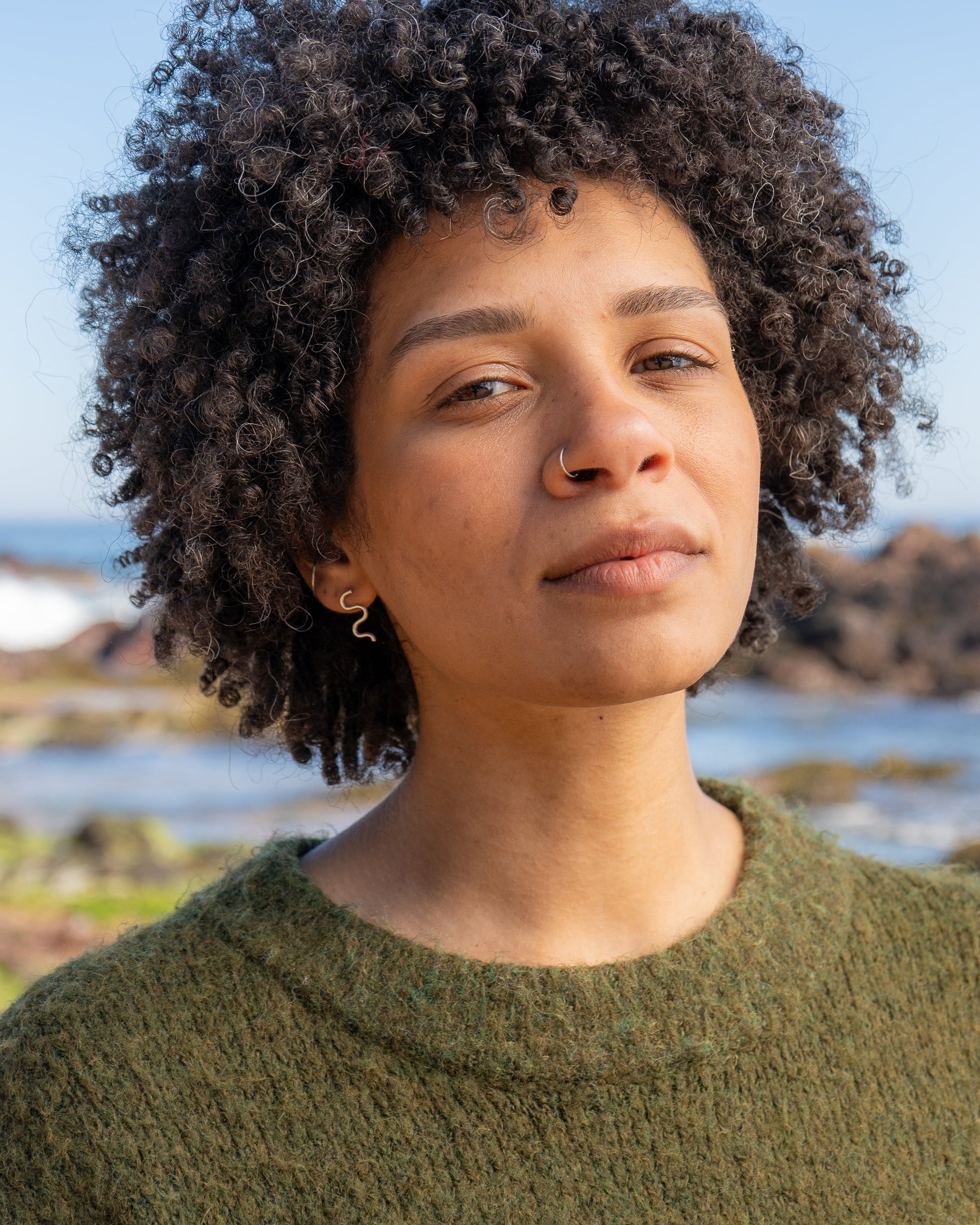 Woman standing on a rocky beach wearing mini wiggle studs with ocean and clear sky in the background