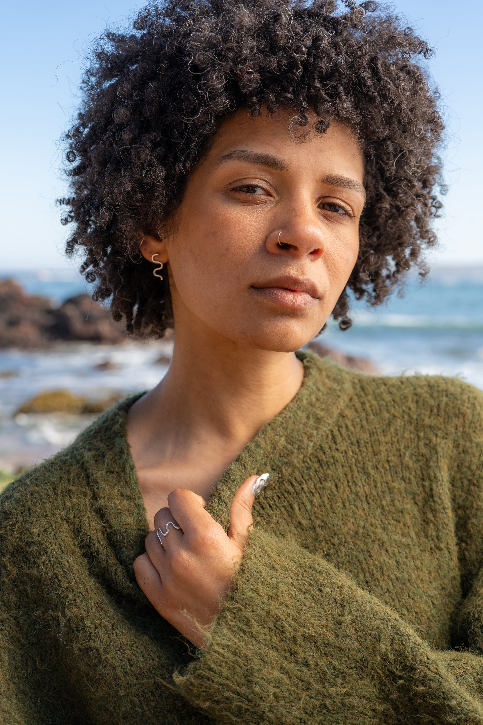 Woman wearing a green sweater wearing mini wiggle studs and wiggle ring with a blurred beach background