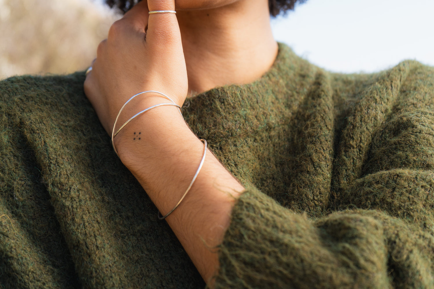 A woman wearing a green jumper with 3 silver bangles on her wrist with blue sky in the background