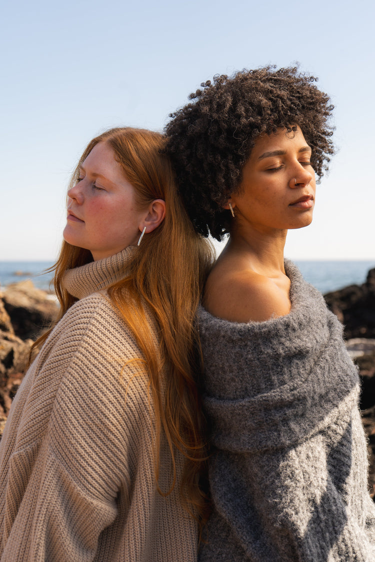 Two women wearing winter jumpers stand back to back wearing long recycled silver earrings on the beach 