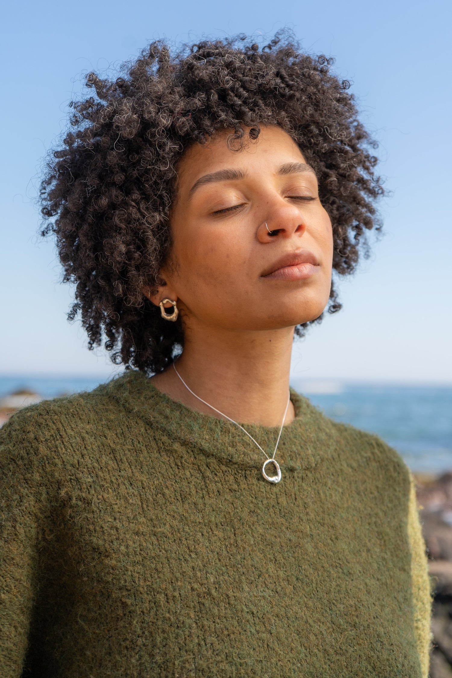 A woman wearing a green jumper has her eyes closed wearing a silver molten necklace and silver molten earrings by the sea
