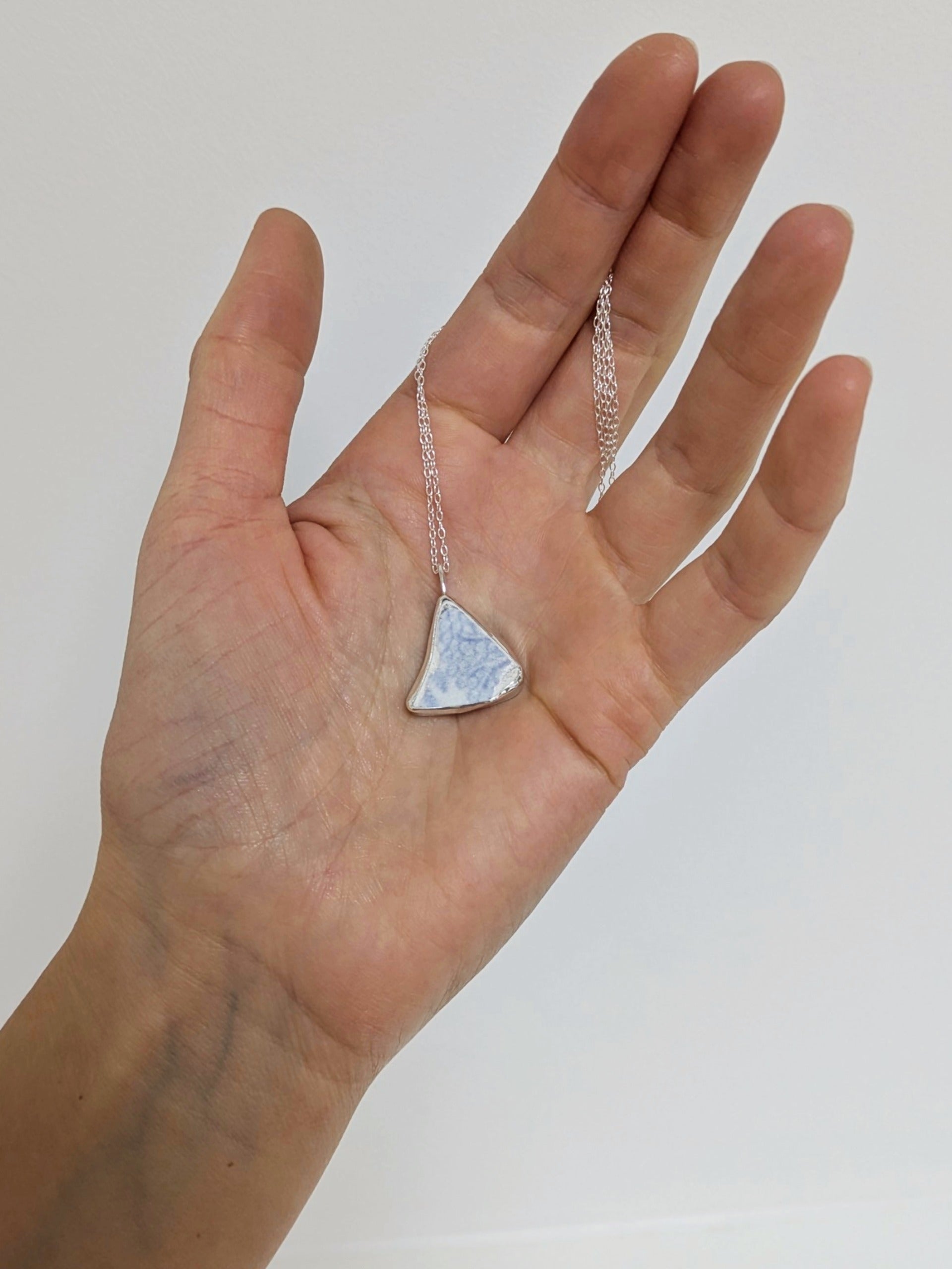Hand holding a silver necklace with a triangular blue floral sea pottery pendant against a white background