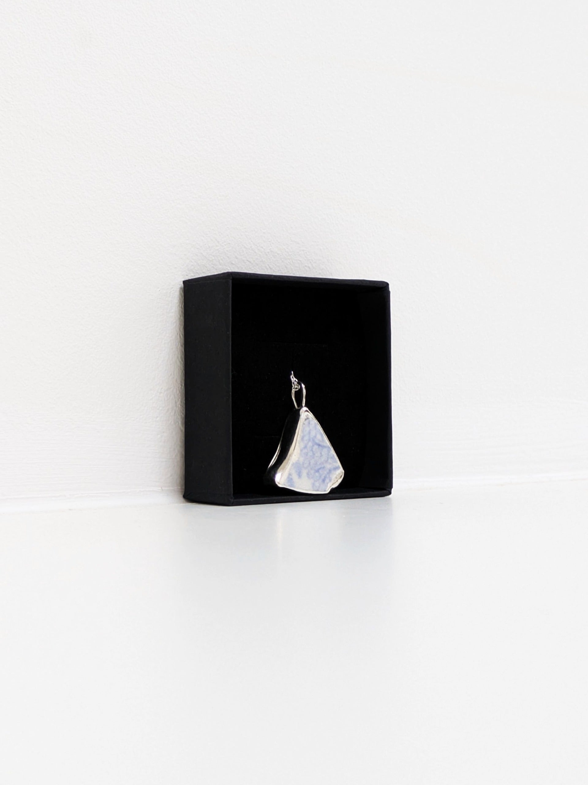Close up angle of a black jewellery display box with a blue floral sea pottery pendant on a white background