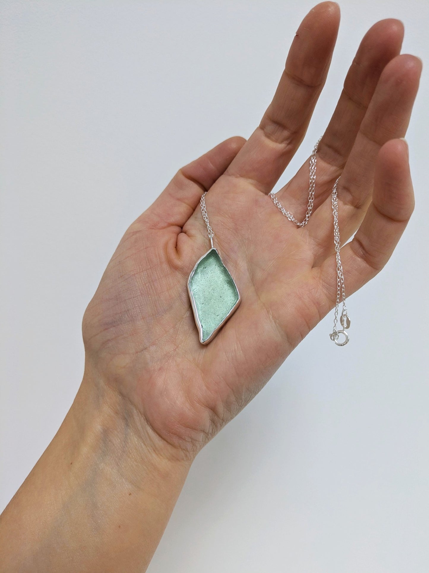 Close up of a hand holding a big turquoise sea glass pendant with a silver chain on a white background.