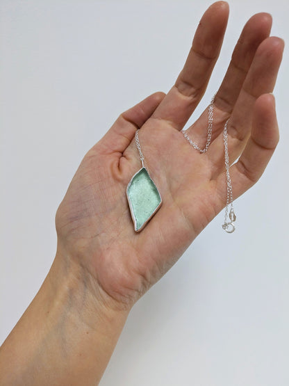 Close up of a hand holding a big turquoise sea glass pendant with a silver chain on a white background.