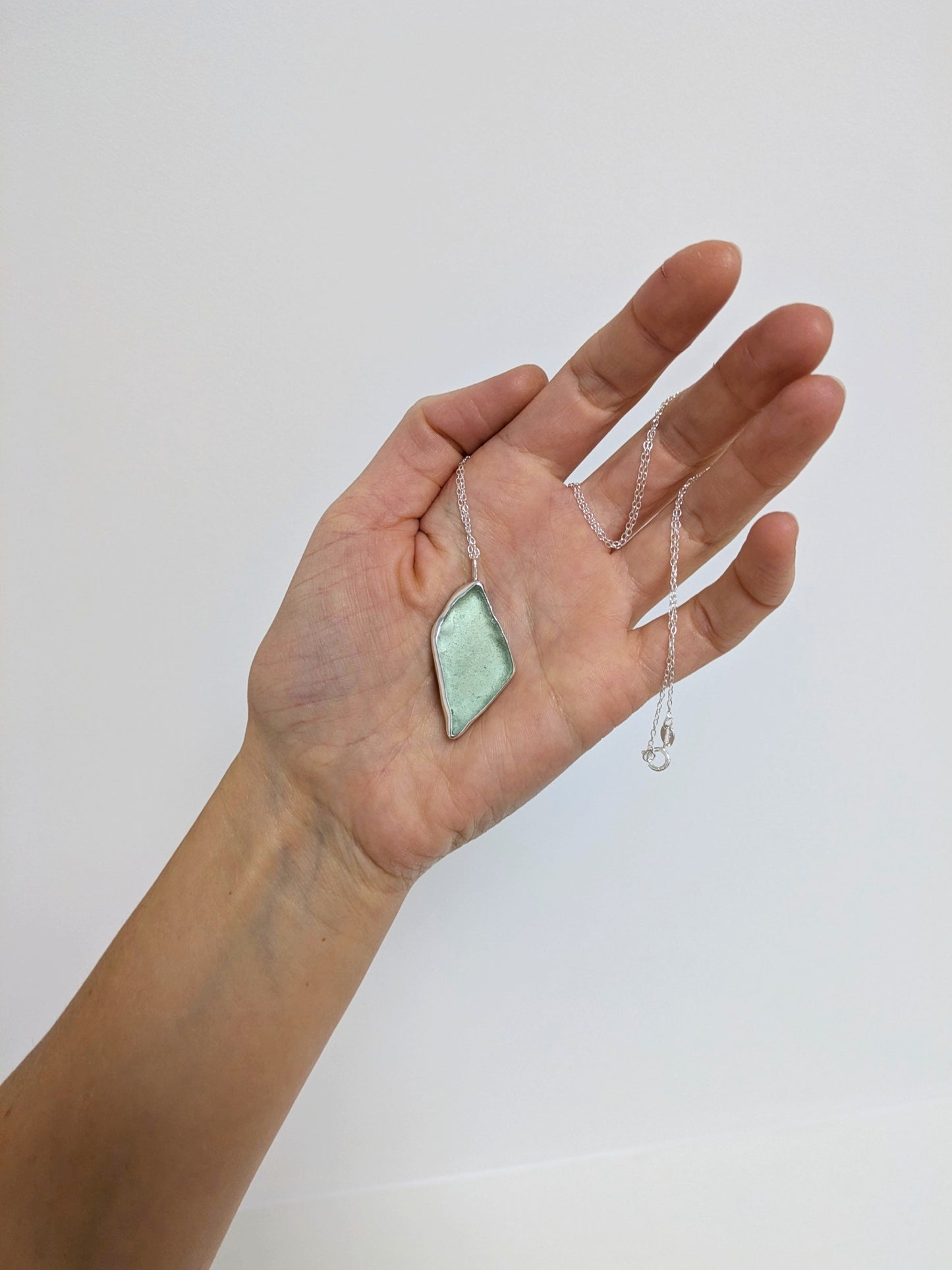 Hand holding a big turquoise sea glass pendant with a silver chain on a white background.