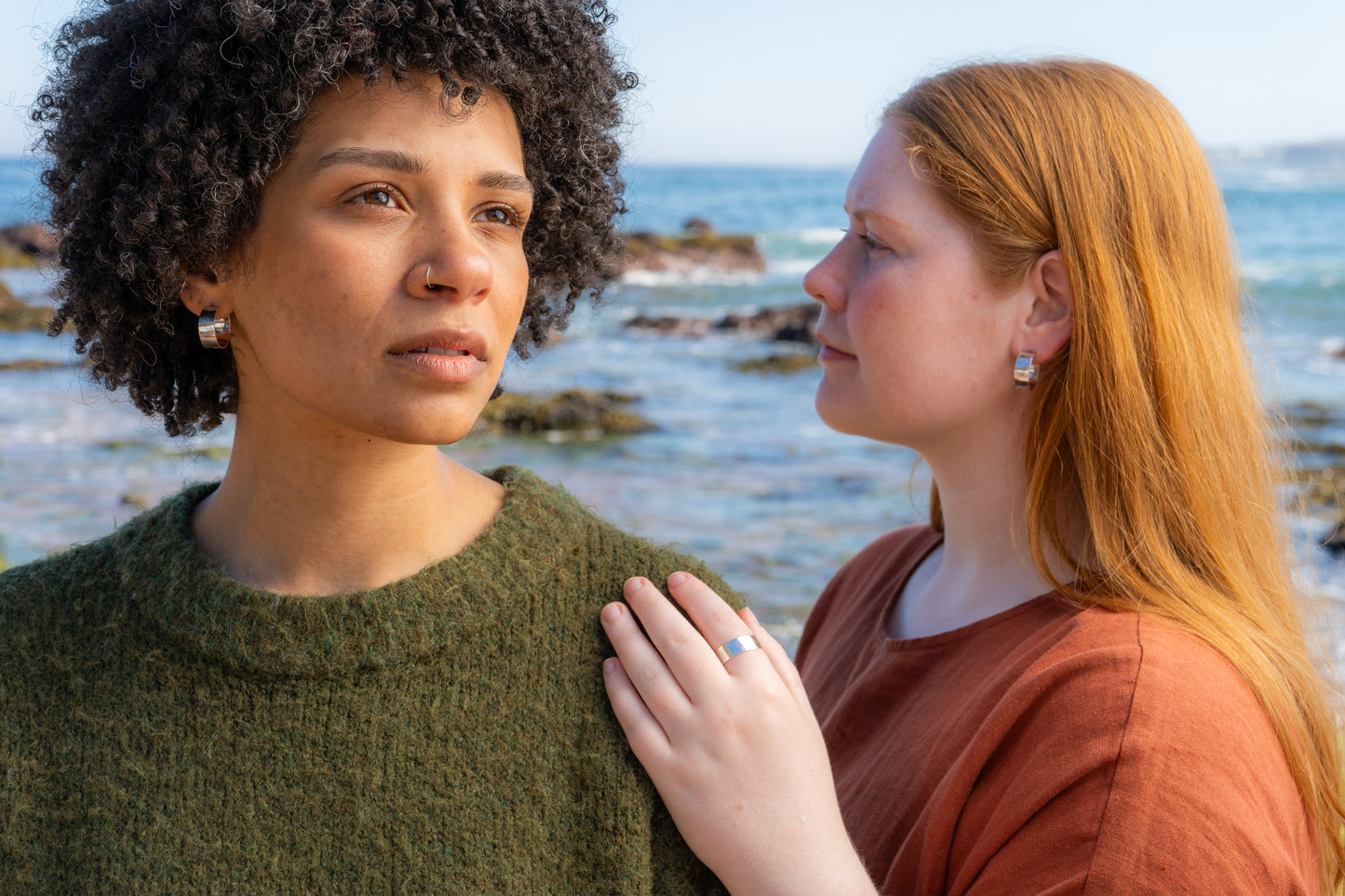 Two women standing close to each other by the ocean wearing hammered hoops