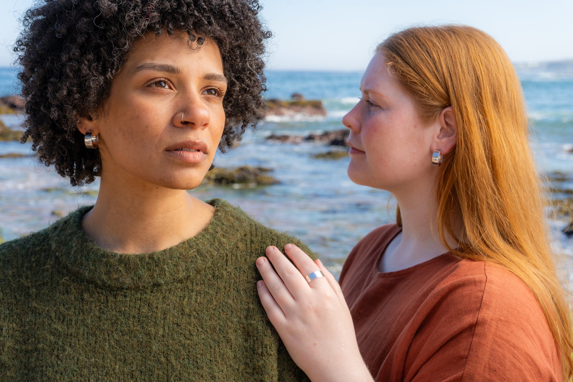 Two women standing close to each other by the ocean wearing hammered hoops