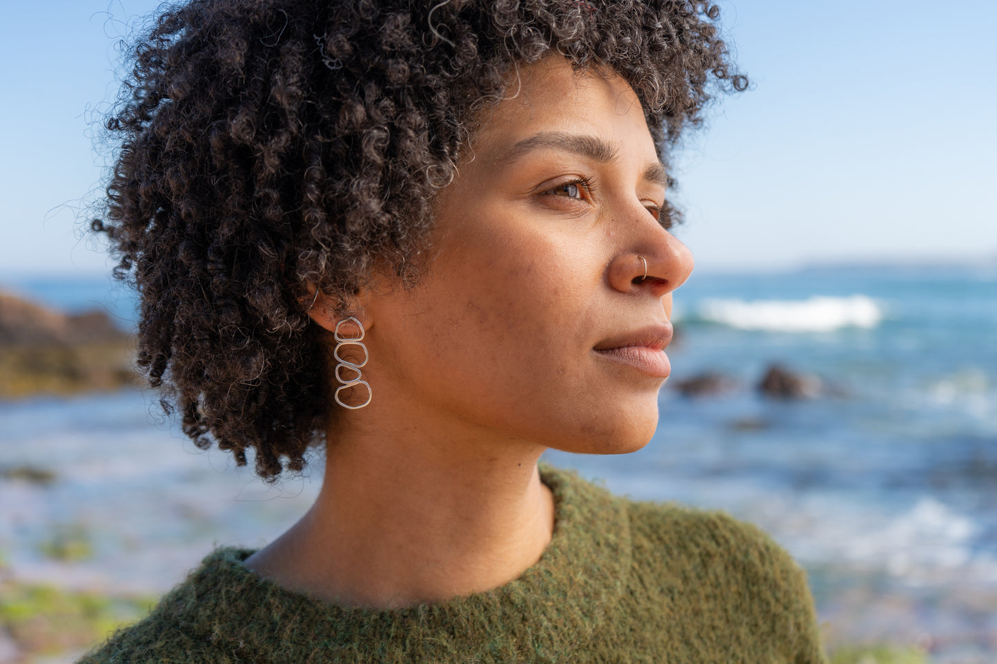Woman with curly hair standing by a coastal landscape wearing chunky stacked pebble earrings
