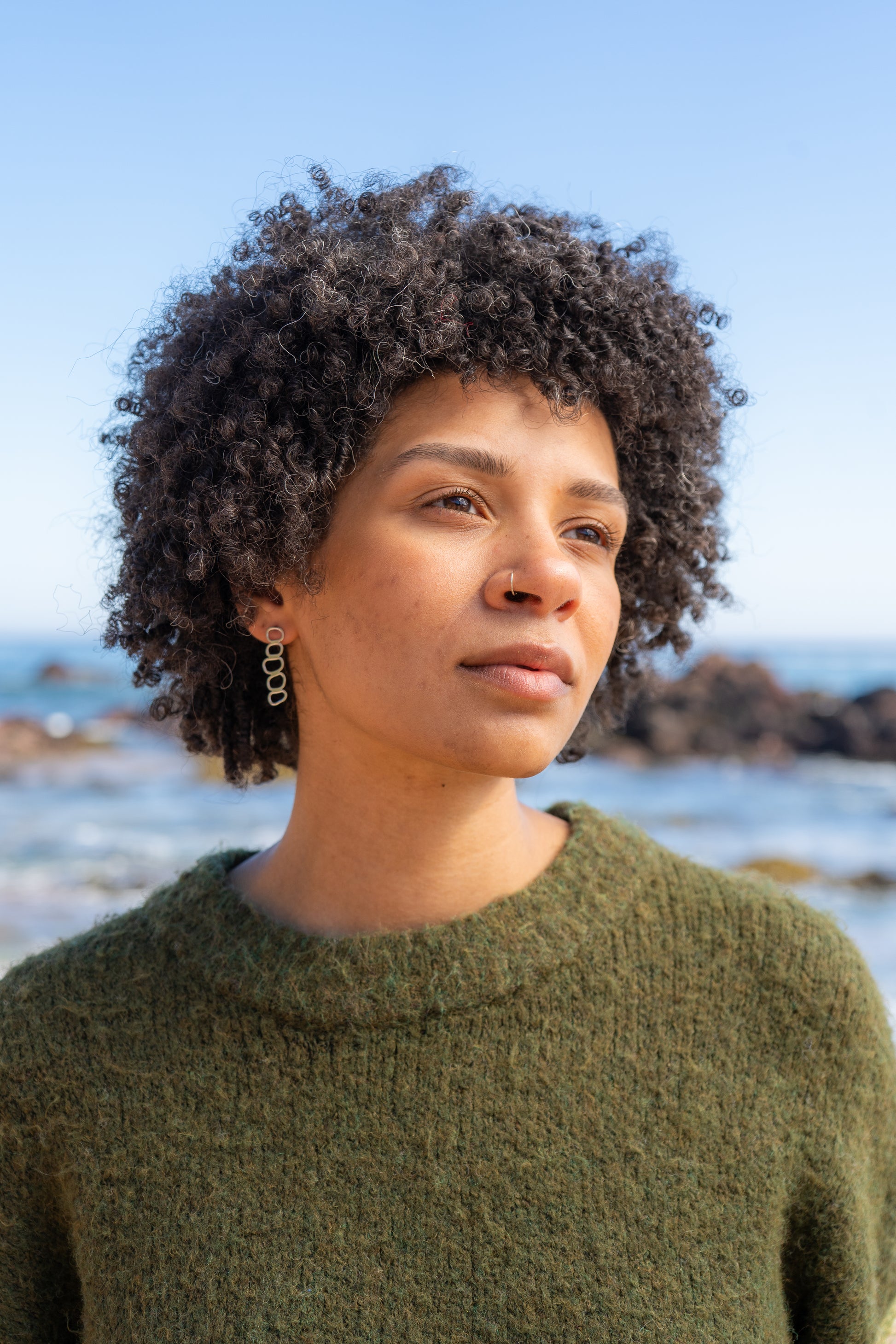 Woman with curly hair wearing a green jumper wear stacked pebble earrings by the ocean