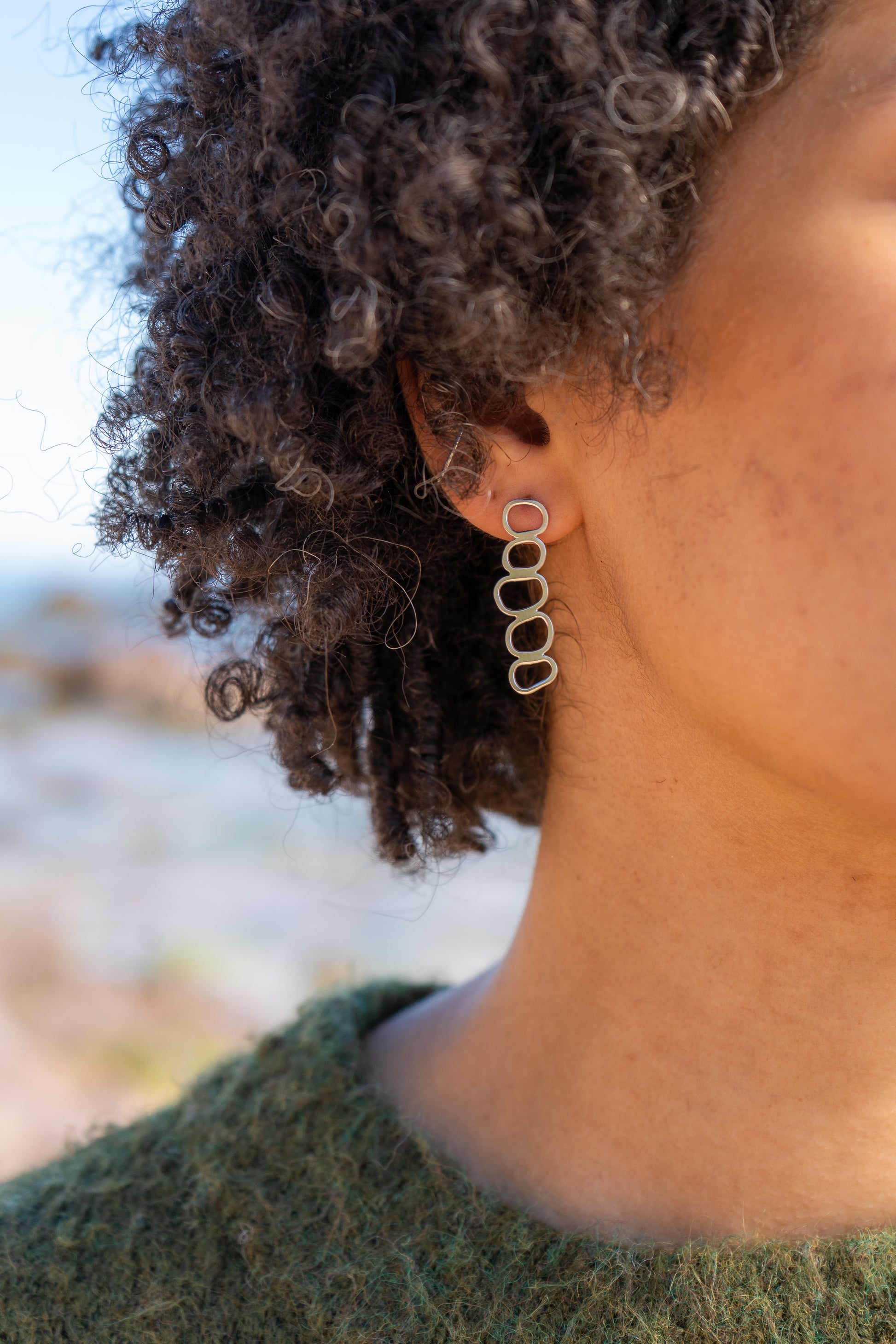 Close-up of a person wearing silver pebble stack earrings with the blurred sea in the background