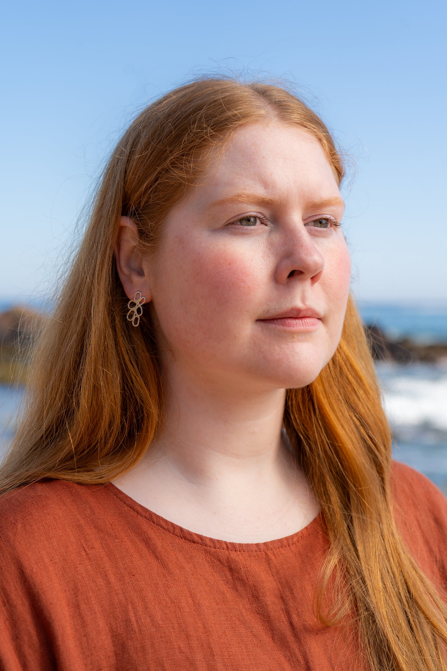 Woman with long red hair wearing an orange dress against a blurred natural background wearing cairn-shaped earrings