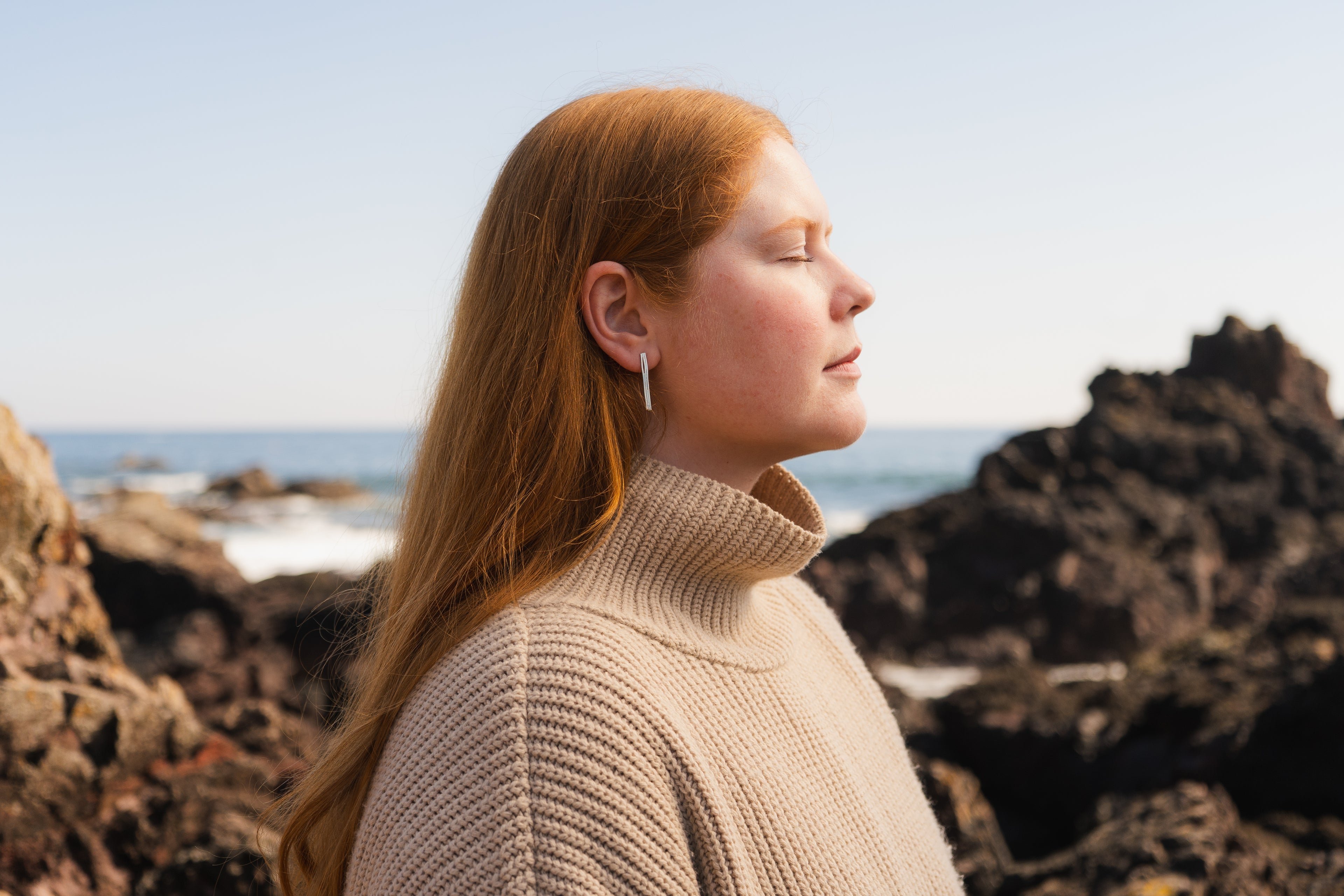 Woman stands on a rocky beach in a brown jumper wearing long earrings.