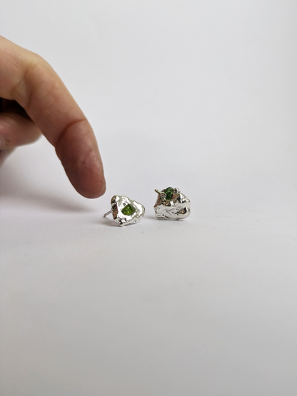 Hand next to a pair of scrap silver earrings with green sea glass shards embedded, on a light grey background.