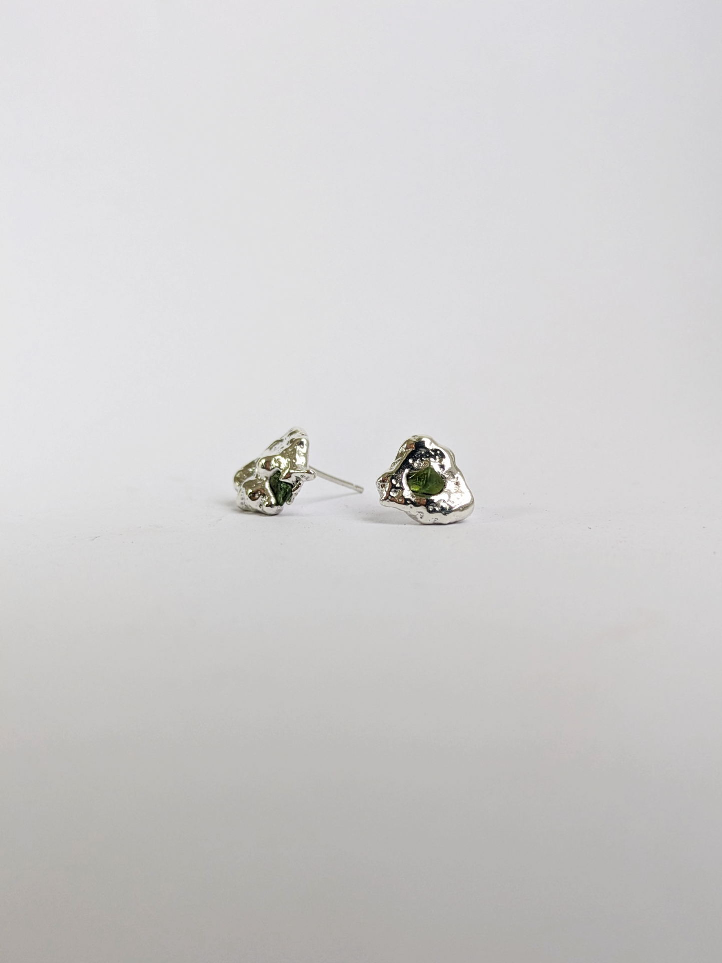 Side view of  a pair of scrap silver earrings with green sea glass shards embedded, on a light grey background.