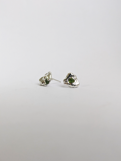 Side view of  a pair of scrap silver earrings with green sea glass shards embedded, on a light grey background.